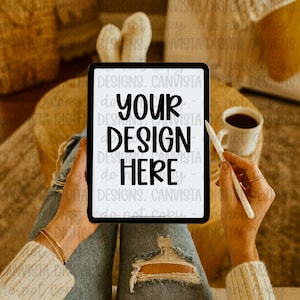 May include: A black tablet displaying the text "YOUR DESIGN HERE" is held by a person wearing a white sweater and ripped blue jeans. A white stylus and a mug of coffee are also visible.
