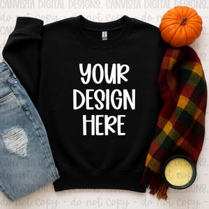 May include: A black crewneck sweatshirt with the text "YOUR DESIGN HERE" in white. The sweatshirt is paired with a pair of blue jeans, a fall-themed scarf, a small pumpkin, and a candle.