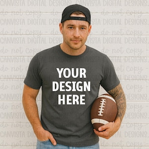 May include: A dark gray t-shirt with the text "YOUR DESIGN HERE" in white. The person in the image is wearing a black baseball cap and holding a brown and white football. The shirt is a crew neck style.