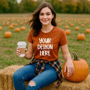Pode incluir: Uma mulher vestindo uma camiseta cor de ferrugem com o texto "YOUR DESIGN HERE" em branco. Ela está sentada em um fardo de feno com uma abóbora e segurando uma xícara de café. Ela está usando jeans azuis e uma camisa xadrez amarrada na cintura.