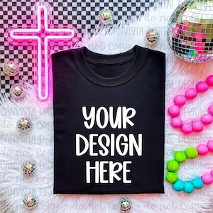 May include: Black t-shirt with the text "YOUR DESIGN HERE" in white. The shirt is surrounded by decorative elements, including a pink neon cross, disco balls, and colorful beads. The background features a checkered pattern.