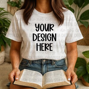 Könnte beinhalten: Weißes T-Shirt mit dem Text "YOUR DESIGN HERE" in Schwarz. Die Person trägt Jeansshorts und hält ein offenes Buch. Das Bild ist eine Lifestyle-Aufnahme, ideal für Mockups.