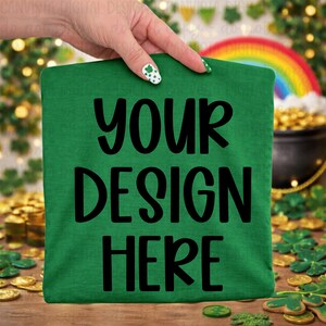 Puede incluir: Una camiseta verde con el texto "YOUR DESIGN HERE" en letras negras grandes. La camiseta está sujeta por una mano con un diseño de uñas con temática de trébol. Un arcoíris y una olla de oro están en el fondo.