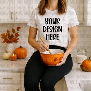 May include: White t-shirt with the text "YOUR DESIGN HERE" in black. The person is holding an orange bowl with a wooden spoon, and the bowl has a fall leaf design. Pumpkins and fall decor are in the background.