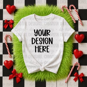 Puede incluir: Una camiseta blanca con el texto "YOUR DESIGN HERE" en negro, anudada a la cintura. La camiseta está rodeada de decoraciones rojas en forma de corazón, lazos rojos y bastones de caramelo sobre una superficie verde y esponjosa, sobre un fondo a cuadros blanco y negro.