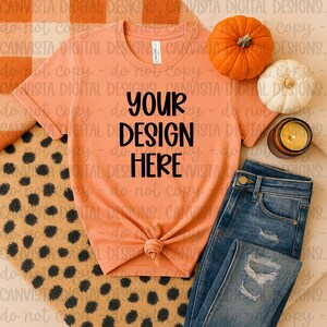 May include: A peach-coloured t-shirt with the text "YOUR DESIGN HERE", knotted at the waist, alongside blue jeans. The image features a small orange pumpkin, a white pumpkin, and a lit candle, suggesting an autumn theme.