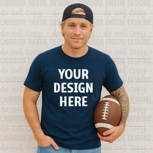 May include: A navy blue t-shirt with the text "YOUR DESIGN HERE" in white. The person in the image is wearing a black baseball cap and holding a brown football. The shirt is short-sleeved.