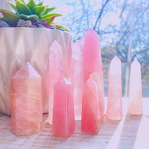 May include: A collection of eight pink rose quartz crystal points, varying in size and shape, arranged on a light wood surface. The crystals are smooth and polished, with a natural, translucent appearance.