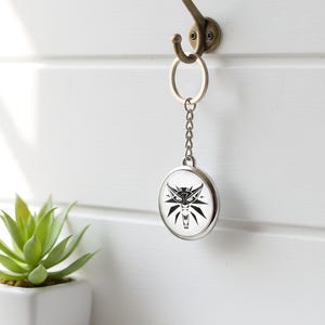 May include: A silver keychain featuring a black and white wolf head design, suspended from a bronze hook on a white wall. The round keychain is attached to a chain. A small potted succulent is on a shelf below.