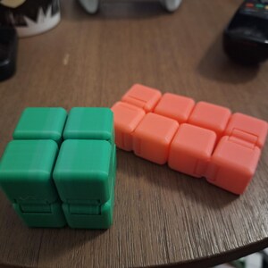 May include: Two fidget cubes are displayed on a wooden surface. One is green, composed of four connected squares. The other is orange, made of eight connected squares. The cubes are designed to be manipulated and provide tactile stimulation.
