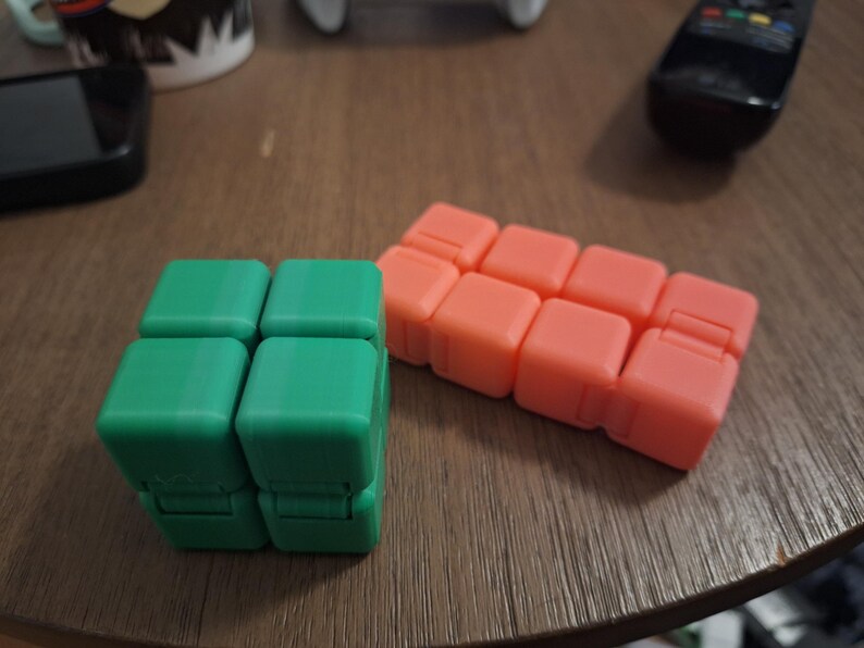 May include: Two fidget cubes are displayed on a wooden surface. One is green, composed of four connected squares. The other is orange, made of eight connected squares. The cubes are designed to be manipulated and provide tactile stimulation.