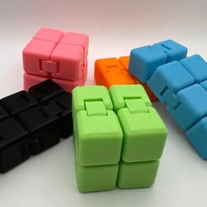 May include: A collection of colorful fidget cubes in various colors, including pink, blue, green, orange, and black. Each cube is composed of smaller, connected blocks that can be manipulated and rotated. The cubes are designed for stress relief and sensory play.