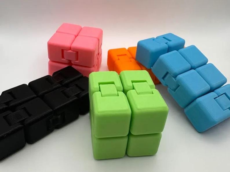 May include: A collection of colorful fidget cubes in various colors, including pink, blue, green, orange, and black. Each cube is composed of smaller, connected blocks that can be manipulated and rotated. The cubes are designed for stress relief and sensory play.