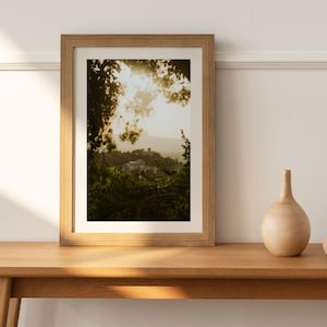 May include: A framed landscape photograph featuring a sunlit scene of a hillside village, viewed through tree branches. The frame is light-colored wood, and a small, light-colored vase sits on the wooden table in front of the frame.