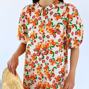 May include: White button-down dress with a vibrant orange fruit and red floral pattern. The dress features short, puffed sleeves and a ruffled hem. A straw sun hat is held in the hand.