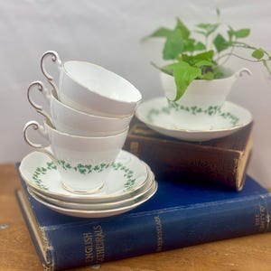 May include: A set of four white teacups and saucers with a green shamrock pattern. The cups are stacked on top of each other and the saucers are placed underneath. The cups and saucers are sitting on top of a stack of books.