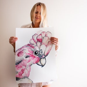 May include: A woman holds a pink and white watercolor painting of a cockatoo bird. The bird's head is turned to the side, and its eye is visible. The painting is signed "Alice" in the bottom right corner.