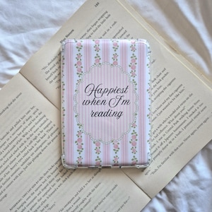 May include: A rectangular case with a pink and white striped design and floral details, inscribed with "Happiest when I'm reading". The case is placed on an open book, with the text visible.