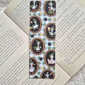 May include: A rectangular bookmark featuring a repeating pattern of white and green gingham squares interspersed with yellow stars and black flowers. The design includes oval frames containing illustrations of white geese wearing witch hats.