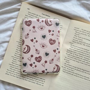 May include: A transparent phone case with a pink backdrop, patterned with hearts, moons, stars, and arrows. The case rests on an open book. The design uses burgundy and black hues. The book's text is visible.