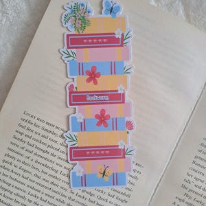 Peut inclure: Un marque-page vertical représentant une pile de livres illustrés en jaune, rose et bleu. Le motif comprend des accents floraux, un papillon bleu, un papillon jaune et une tranche rouge avec le texte Bookworm imprimé en lettres blanches.