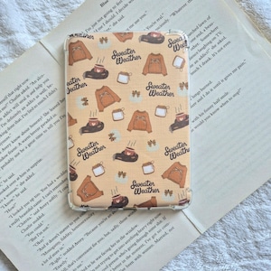 May include: A clear protective case for an electronic device featuring a repeating pattern on a light orange background. The design includes illustrations of brown sweaters, mugs of hot beverages, and the text Sweater Weather in black script.