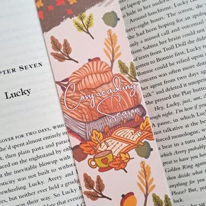 Peut inclure: Un marque-page rose sur le thème de la lecture confortable. Le marque-page présente une illustration dessinée à la main d'une personne lisant un livre avec une tasse de café et une couverture. Le texte "Cozy Reading Season" est écrit en cursive. Le marque-page est décoré de feuilles d'automne et de glands.