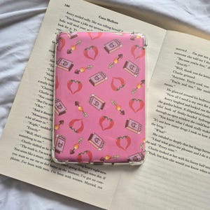 May include: A pink phone case with a repeating pattern of red chili peppers, books, and arrows. The case is rectangular and appears to be made of a clear, protective material. The case is resting on an open book.