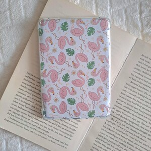 May include: A clear phone case with a repeating pattern of pink flamingos, flowers, and green leaves on a white backdrop. The rectangular case is designed to protect a device, possibly a tablet or e-reader.