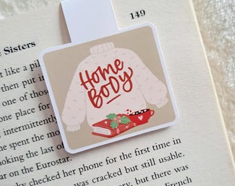 May include: A square magnetic bookmark with a beige background featuring a white knit sweater illustration. The text Home Body is written in red script. Below the sweater is a red book with green holly leaves and a small red mug with white dots.