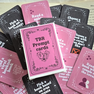 May include: A selection of TBR Prompt Cards in pink and black. The cards have prompts for book choices, with decorative borders and the text "TBR Prompt Cards" on the box. The cards are scattered on a vintage book page.