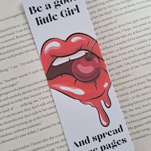 May include: A bookmark with a red, white and black design. The bookmark features a red, cartoon-style mouth with a cherry in it. The text "Be a good little Girl" is printed on the top half of the bookmark. The text "And spread those pages" is printed on the bottom half of the bookmark.