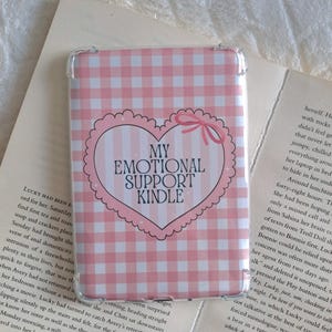 Peut inclure: Une coque Kindle à carreaux roses et blancs avec le texte "MY EMOTIONAL SUPPORT KINDLE" à l'intérieur d'un cœur. L'étui est posé sur un livre ouvert.