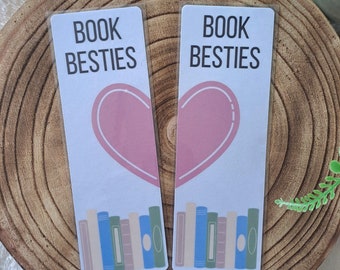 May include: Two white bookmarks with the text "Book Besties" and a pink heart design. Each bookmark features a stack of colourful books at the bottom.