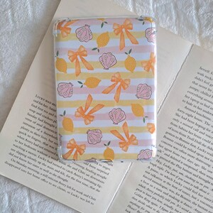 May include: Transparent phone case with a repeating pattern of orange bows, pink seashells, and yellow lemons on a striped pink and yellow background. The case is designed for a smartphone and offers protection.