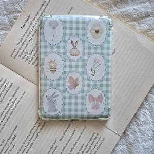 May include: A clear phone case with a light green gingham pattern and oval illustrations of a dandelion, bunny, bee, flower, butterfly, and bow. The case is resting on an open book.
