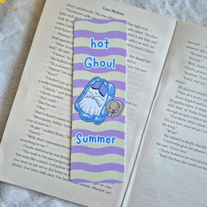 May include: A rectangular bookmark with a wavy purple and light yellow striped background. The text "hot Ghoul Summer" is printed in blue. A cartoon illustration shows a white ghost relaxing in a blue inflatable pool with a sun hat nearby.