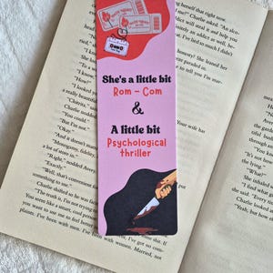 May include: A pink bookmark with the text "She's a little bit Rom-Com & A little bit Psychological thriller." The bookmark features illustrations of movie tickets, a cassette tape, and a knife with blood.