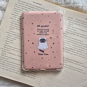 May include: A rectangular e-reader case with a clear plastic frame and a matte pink cover. The cover features a central illustration of a cat wearing a white ghost sheet, surrounded by black dots and the text: Oh goodie! You're trying to talk to me when I'm reading again. How fun.