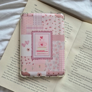 May include: A pink patchwork phone case with a clear border, featuring floral, cherry, and heart patterns. The centre has a postage stamp design with a teacup and hearts. The case is resting on an open book.
