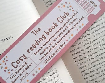 May include: A pink bookmark with a ticket design. It features the text "The Cosy Reading Book Club" and "Admit One". The bookmark has illustrations of autumn leaves and the text "Relax in front of the roaring open fire whilst reading your favourite comfort books. Free hot drinks & cake throughout your stay! Lifetime member."