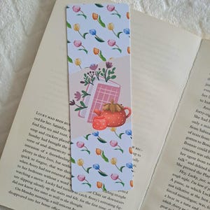 May include: A pink and white bookmark with a floral pattern. The bookmark features a teapot with a strawberry on top, a book, and a bouquet of flowers. The bookmark is ideal for adding a touch of whimsy to your reading experience.