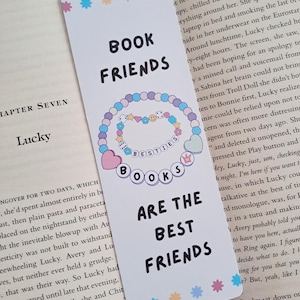 May include: A pink and white bookmark with a colourful illustration of a book with a chain of hearts, stars, and dice. The text "Book Friends" is at the top of the bookmark and the text "Besties Books Are The Best Friends" is at the bottom.