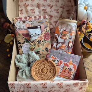May include: A gift box containing a glass tumbler with a bamboo lid and straw featuring autumn graphics, a floral envelope with a bookmark, a sage green scrunchie, a brown crocheted pumpkin coaster, and a card with the text My Autumn is All Booked.