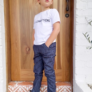 Puede incluir: Una persona joven lleva una camiseta blanca con el texto "The Connections" impreso en la parte delantera. Lleva pantalones de rayas azules oscuros y zapatillas negras. La persona está de pie frente a una puerta de madera con una pared de ladrillo blanco a la derecha.