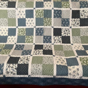May include: A patchwork quilt featuring a grid of square fabric blocks in shades of navy blue, sage green, and white. The blocks display various patterns including floral prints, geometric grids, and stripes, all separated by white sashing.