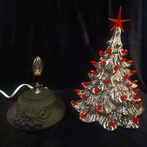 May include: A ceramic Christmas tree with red lights and a red star on top, next to a black base with a light bulb. The tree is silver and black, with red lights. The base has a holly design.