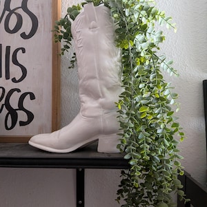May include: A white ceramic cowboy boot vase filled with green leafy plants. The boot is on a black shelf, with a wooden sign in the background. The sign has black text that reads "Bless this mess."