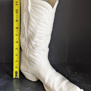 May include: A white ceramic cowboy boot, approximately 12 inches tall, with detailed stitching and a pointed toe. The boot is unpainted and ready for customization. A yellow measuring tape is placed next to the boot.
