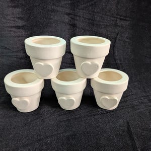 May include: Five small, off-white ceramic flower pots. Each pot has a heart-shaped embellishment on the front. The pots are arranged on a dark background, showcasing their simple, yet charming design. Ideal for small plants or succulents.